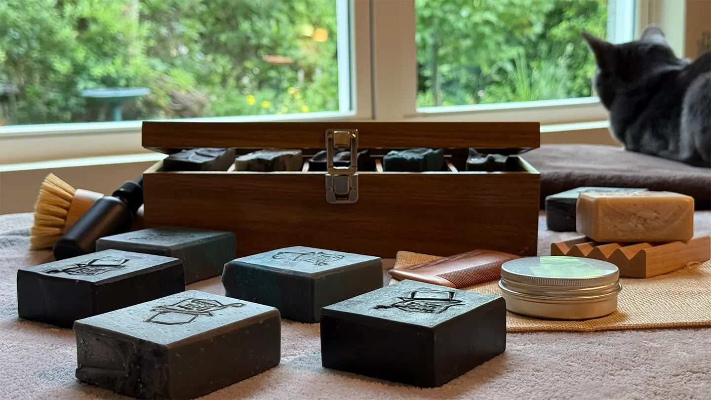 Don Gato Soap Co. The Gentleman Collection — five handcrafted artisan soap bars with activated charcoal in shades of black, gray, and deep teal, displayed with a wooden gentleman's grooming box, shaving brush, wooden comb, and beard balm tin, on a soft fabric surface with a window view of green foliage in the background.