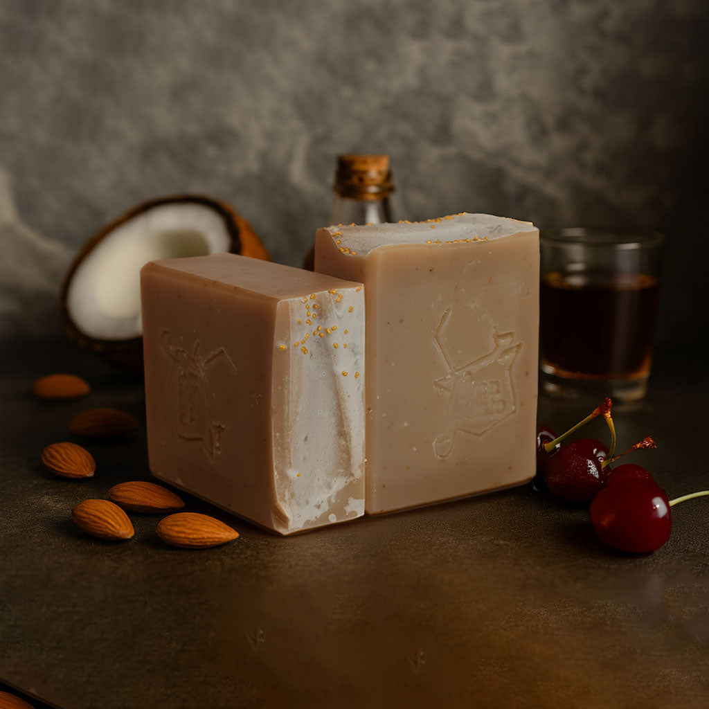 Almond Artisan Soap bars in tan and white with gold speckles, styled with almonds, cherries, coconut, and amber drink on a dark surface.